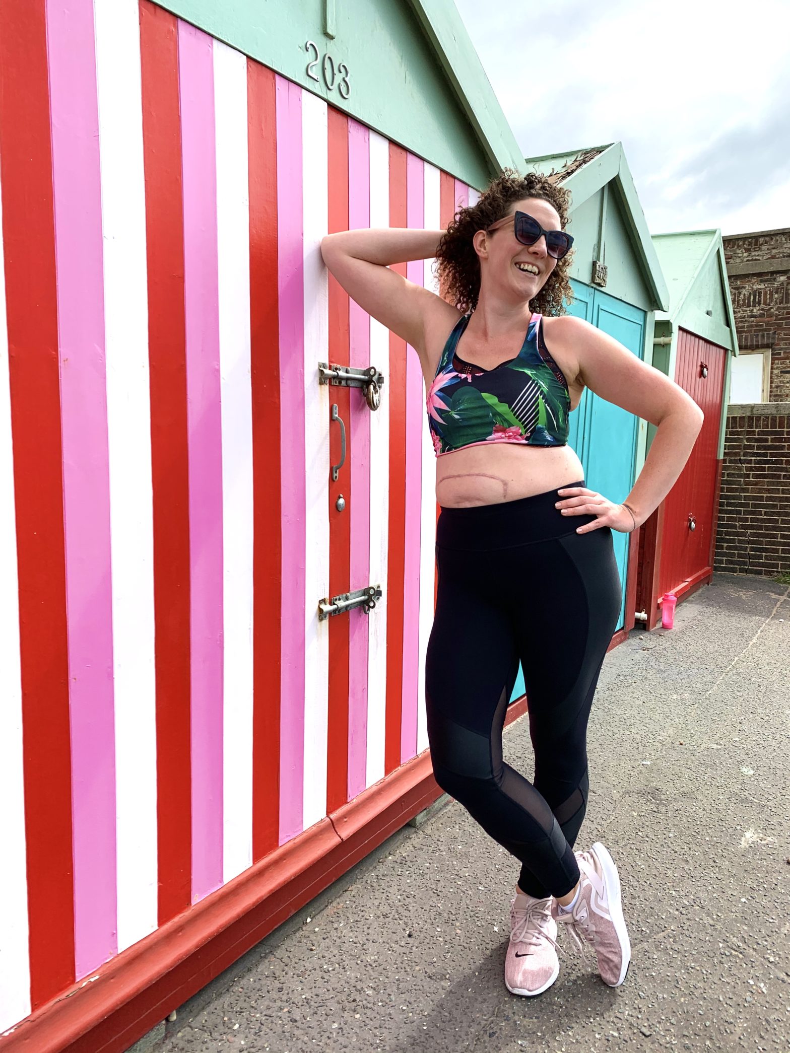 Personal Trainer Becky Hughes leans against a beach hut, smiling with sunglasses on