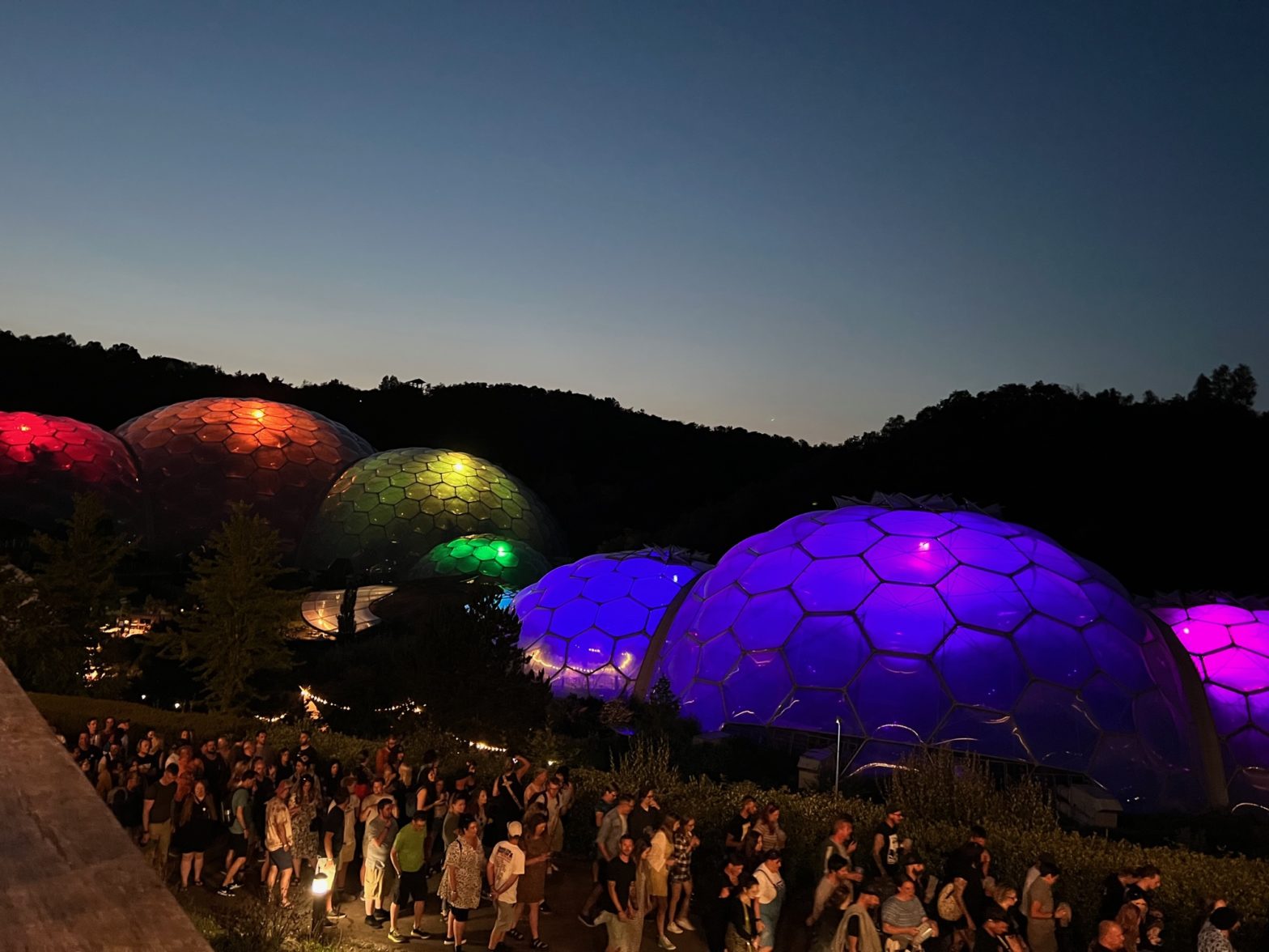 Dusk at Eden Project, Cornwall, looking over the biome domes, each of which is lit up in a colour of the rainbow (left to right, red to violet)