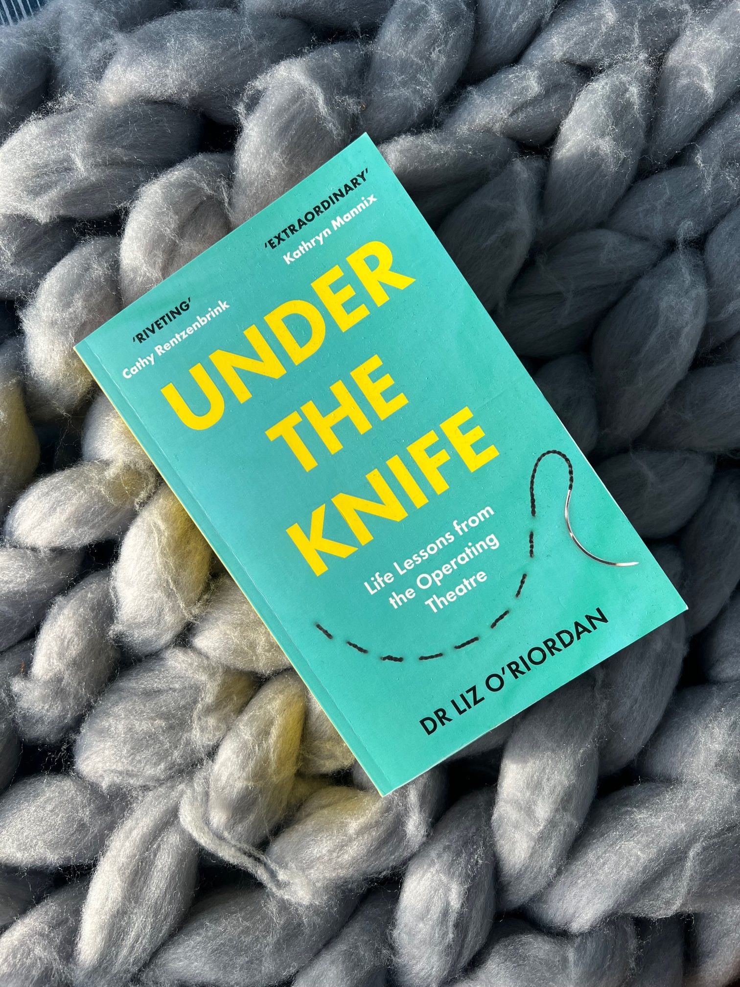 Paperback book "Under the Knife" by Liz O'Riordan sits on a chunky knitted blanket