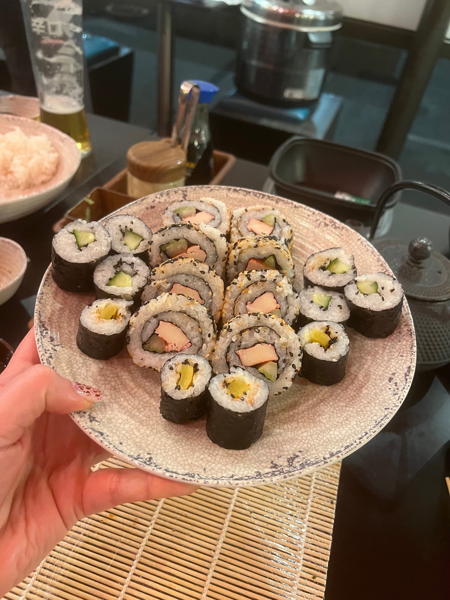 White hand holds a plate of sushi roll portions - maki and California rolls; table is visible beneath the plate, photo is taken indoors