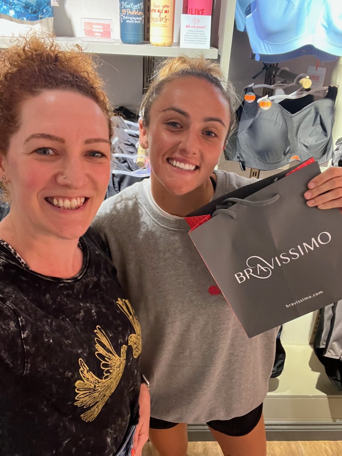 Selfie of two White women posing in front of a display of sports bras. Both are smiling at the camera, and one holds up a "Bravissimo" carrier bag