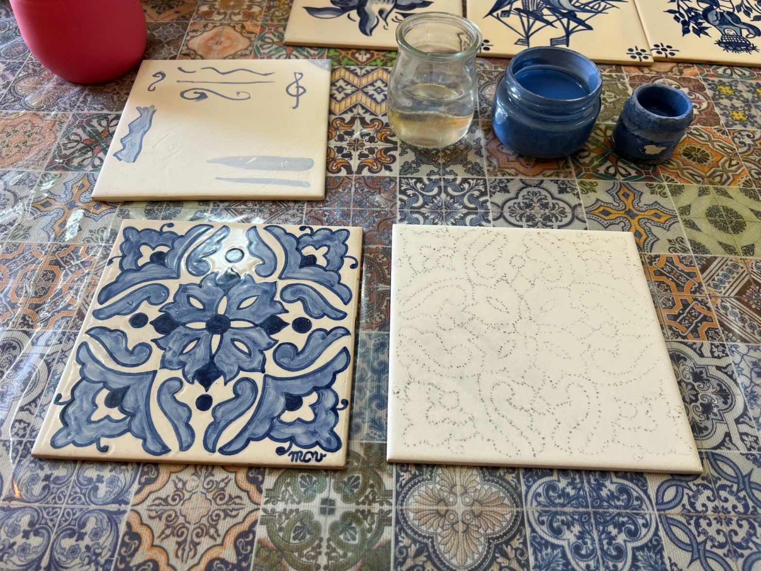 Three ceramic tiles at various stages of design completion on a colourful tabletop. In the background are two small paint pots and a water jar