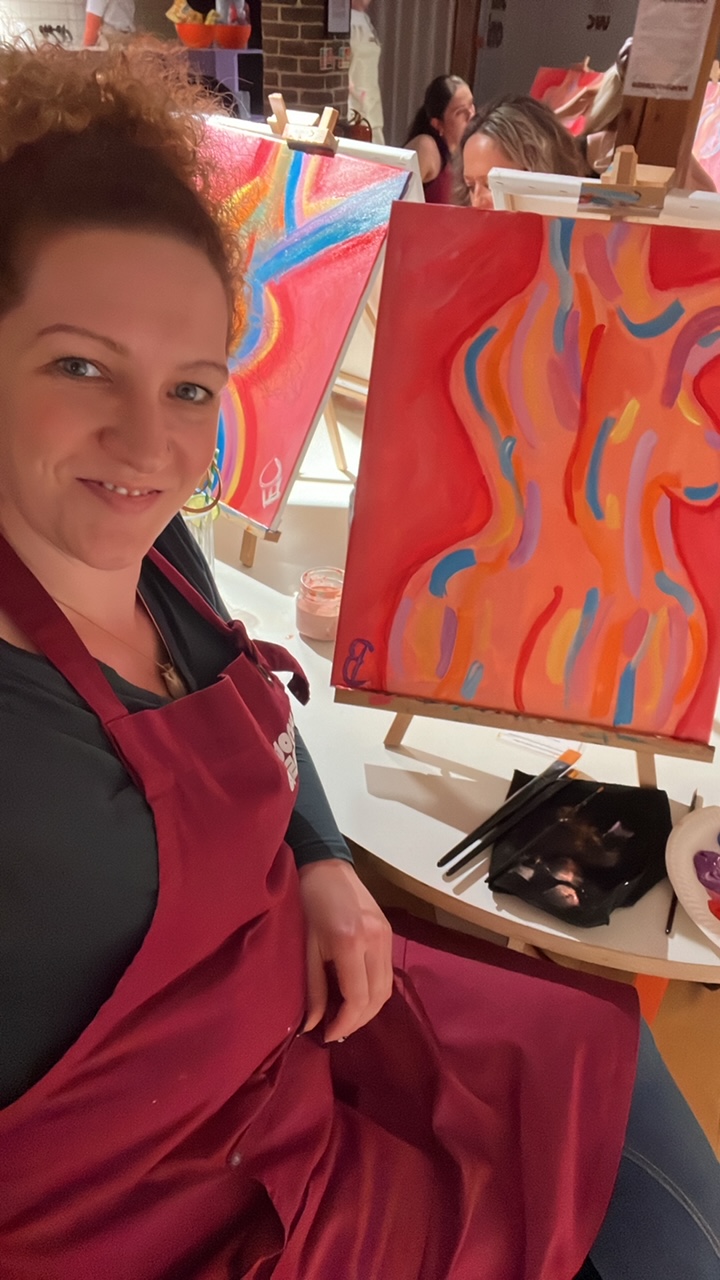 Selfie of White woman seated at indoor art class. She is wearing a dark red apron and her curly hair is tied back. Next to her is a painting she has completed - a stylised female nude