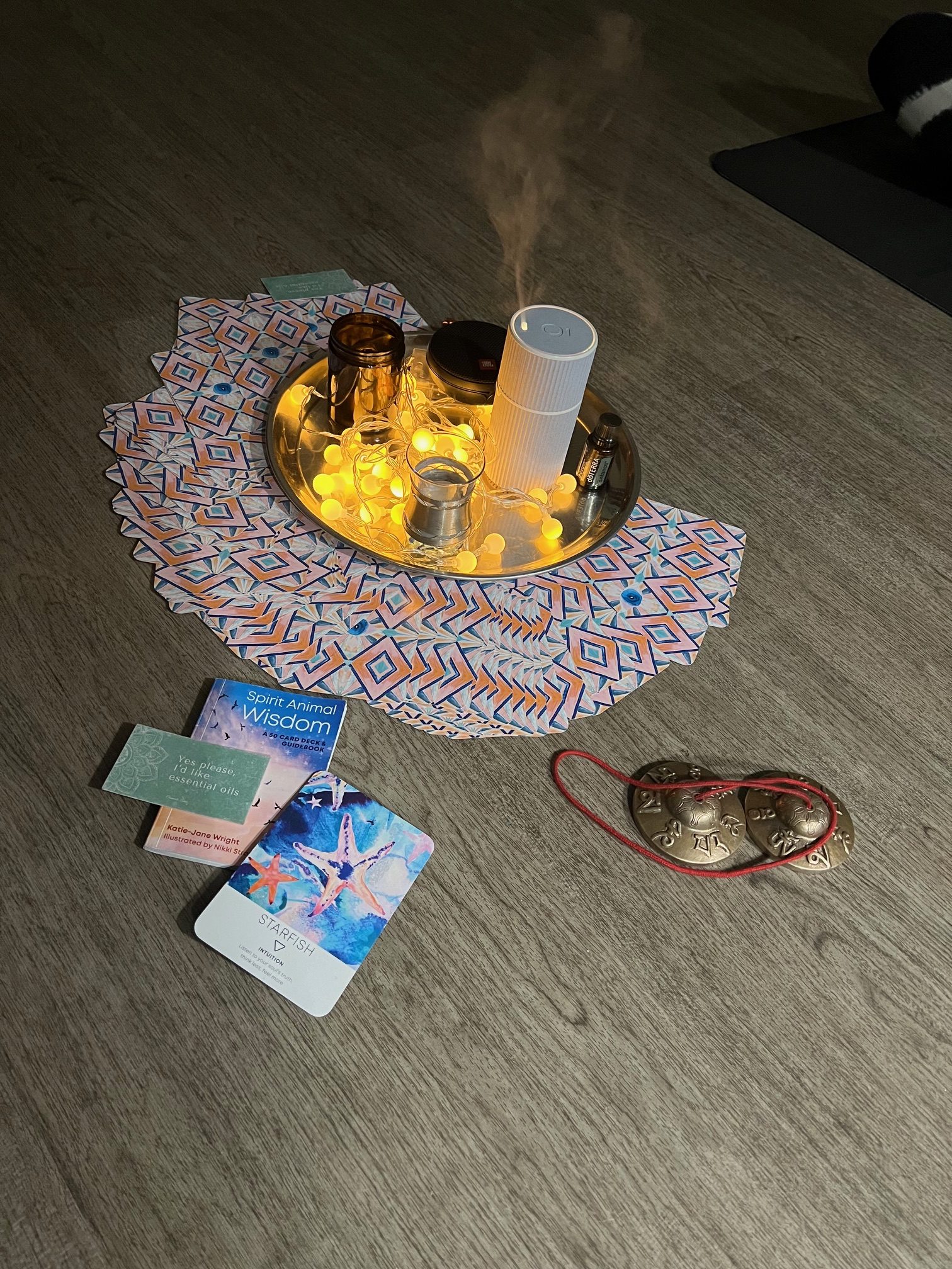 Collection of objects on the floor at a yoga class, representing a focus point: round metal tray contains a string of fairy lights, plus an elecrtic diffuser and candles. Around it is spread a deck of cards, face down. Next to these are a pair of flat bells, plus a selected oracle card and book