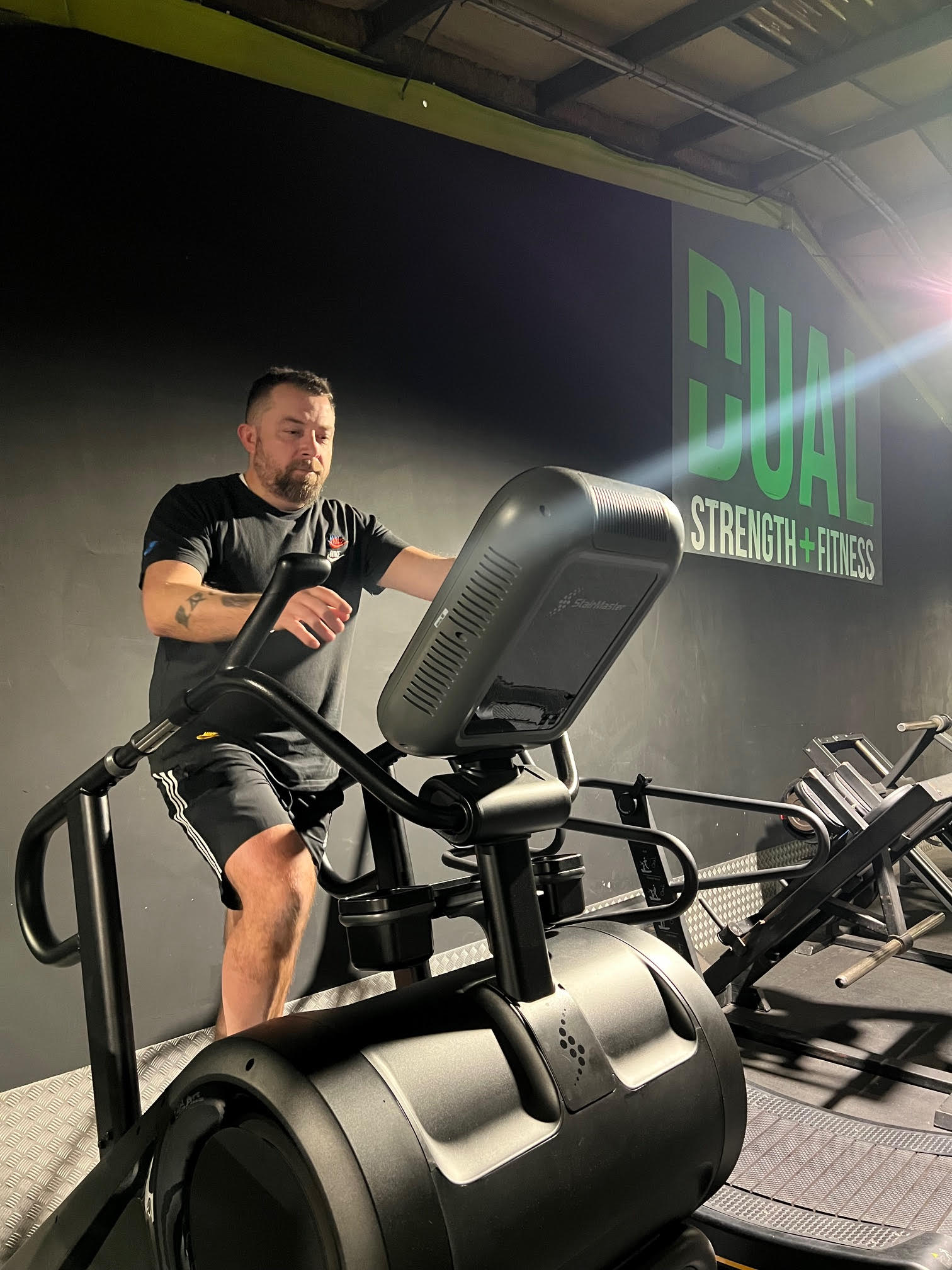 Man wearing black t-shirt and shorts trains on a stair climber indoors at Dual Strength + Fitness gym