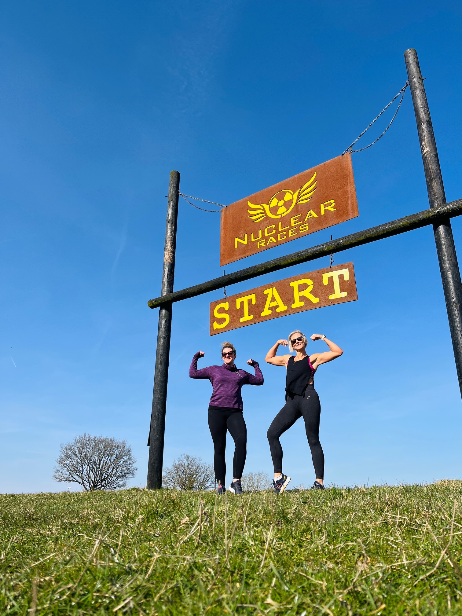 Two white women stand in a sunny field beneath a sign reading "Nuclear Races Start", both are smiling and stand in a double biceps pose