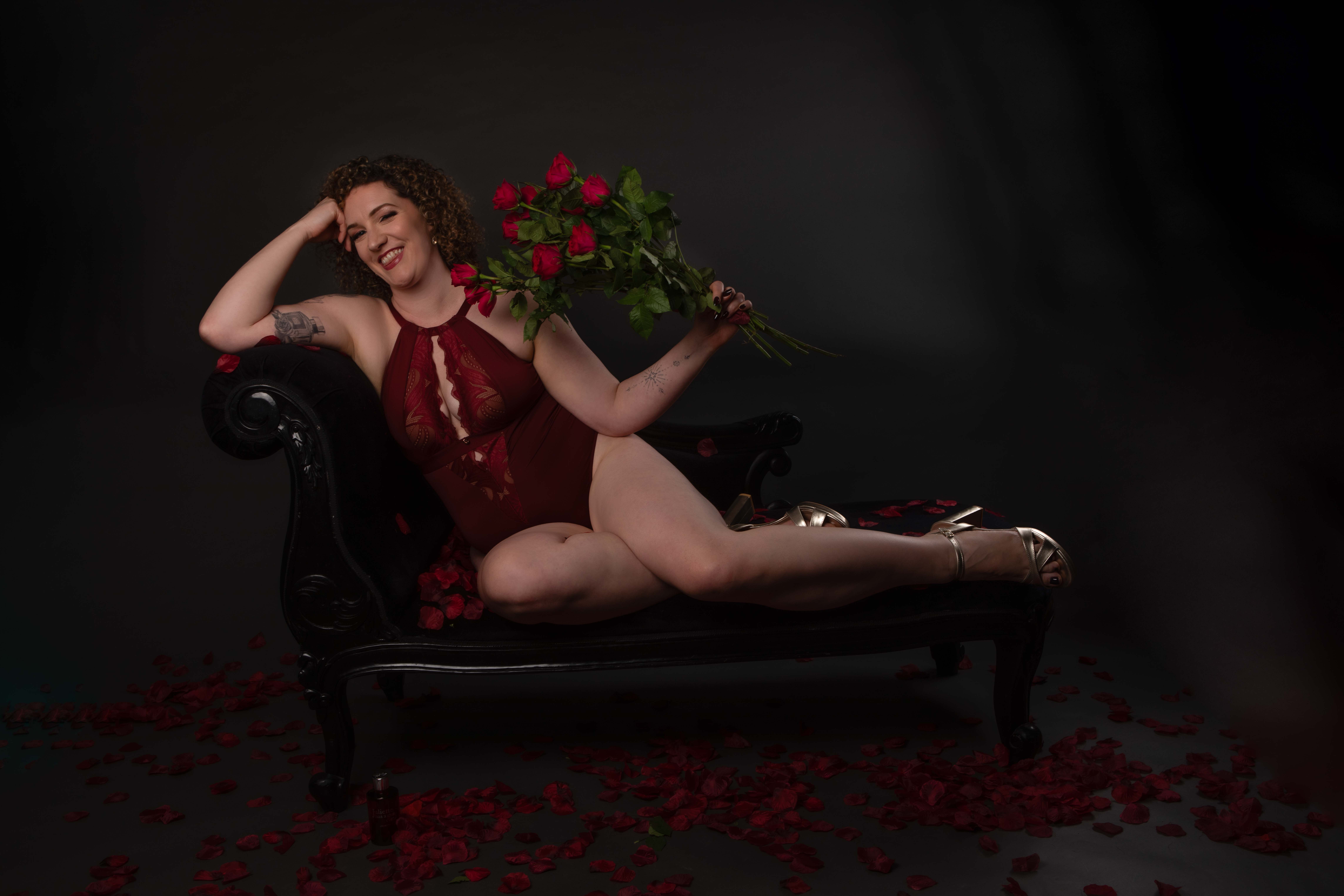 Curly-haired White woman sits on a chaise lounge, one elbow on the back of it, feet up on the seat beside her, holding a bunch of red roses. She wears an oxblood lace bodysuit