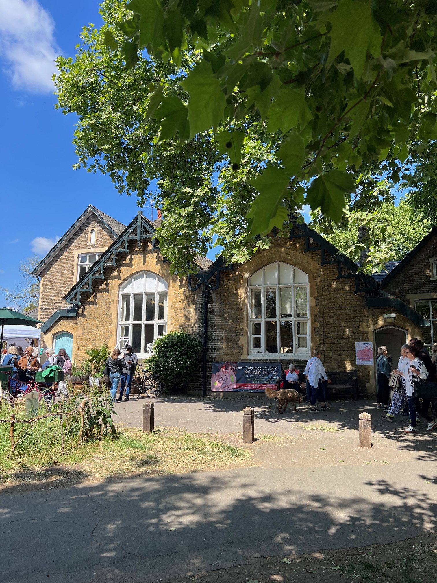 Exterior view of Barnes Fragrance Fair venue - there's a leafy branch in the foreground and blue sky in the background. The event is clearly busy with lots of people milling around