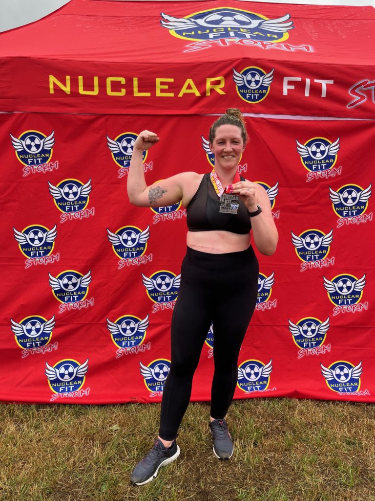 Becky, a White woman, stands in front of a red Nuclear Fit brand wall outside. She wears a black crop top, trainers, and leggings, and holds her finisher medal up in her left hand, with her right arm flexing the bicep