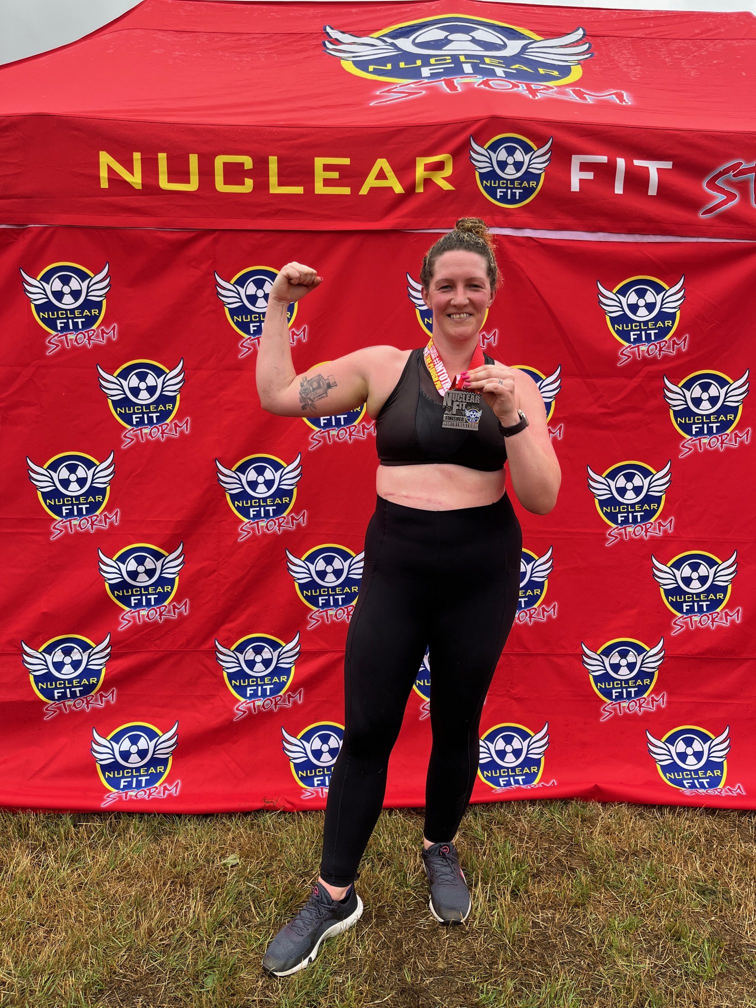 Becky, a White woman, stands in front of a red Nuclear Fit brand wall outside. She wears a black crop top, trainers, and leggings, and holds her finisher medal up in her left hand, with her right arm flexing the bicep