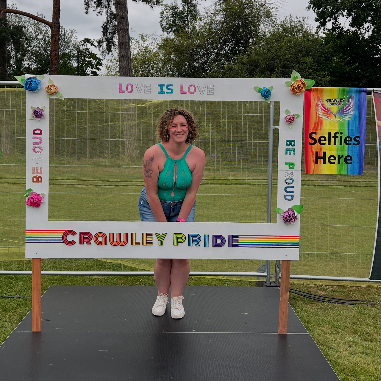 Becky - a White woman - poses in a Crawley Pride selfie frame. She is leaning towards the camera and smiling