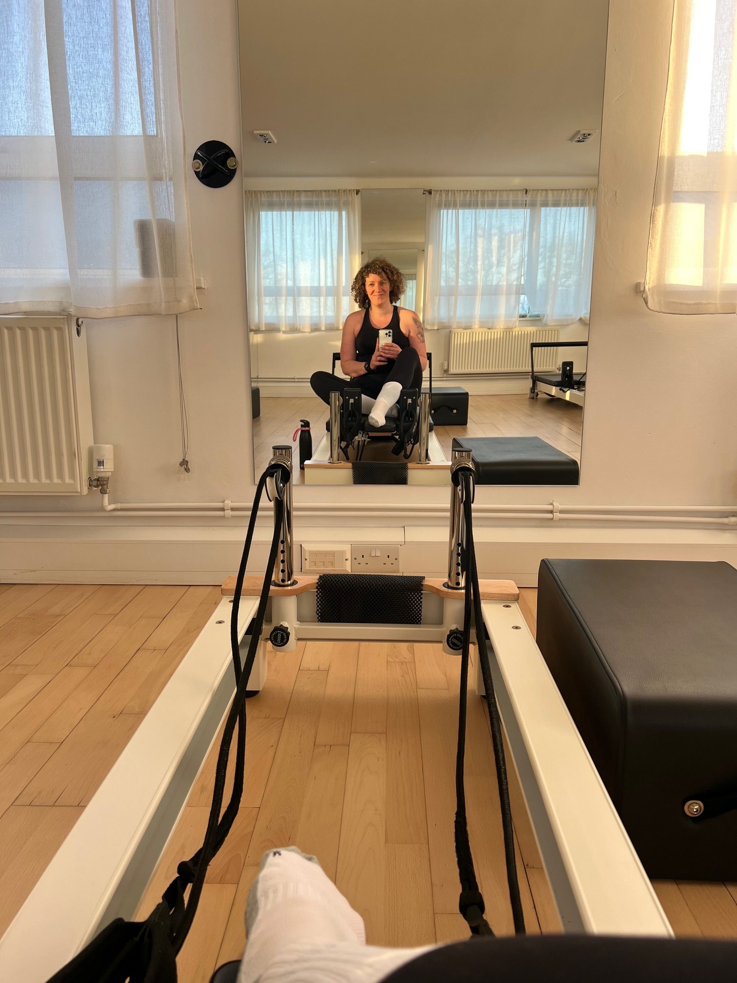 Becky, a White woman, takes a mirror selfie whilst sat on a reformer carriage in a Pilates studio