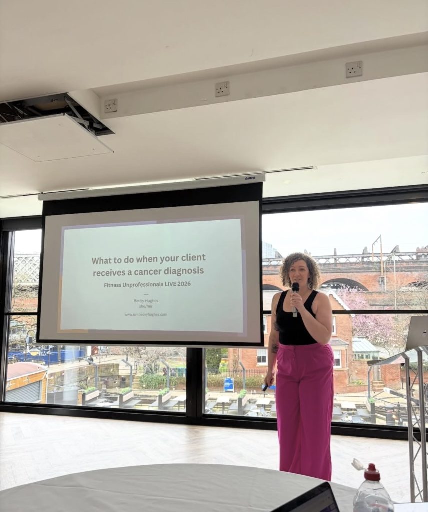 Becky, a curly-haired White woman, stands in front of a screen displaying the title of her talk, "What to do when your client receives a cancer diagnosis". She is holding a microphone, and wears a black vest top and a pair of bright pink tailored trousers