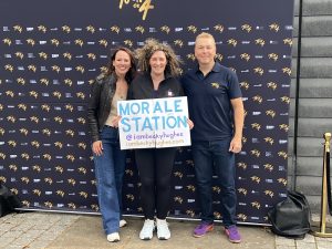 Lady Sarra Hoy, Becky, and Sir Chris Hoy pose with Becky's "Morale Station" sign in front of a Tour de 4 brand wall