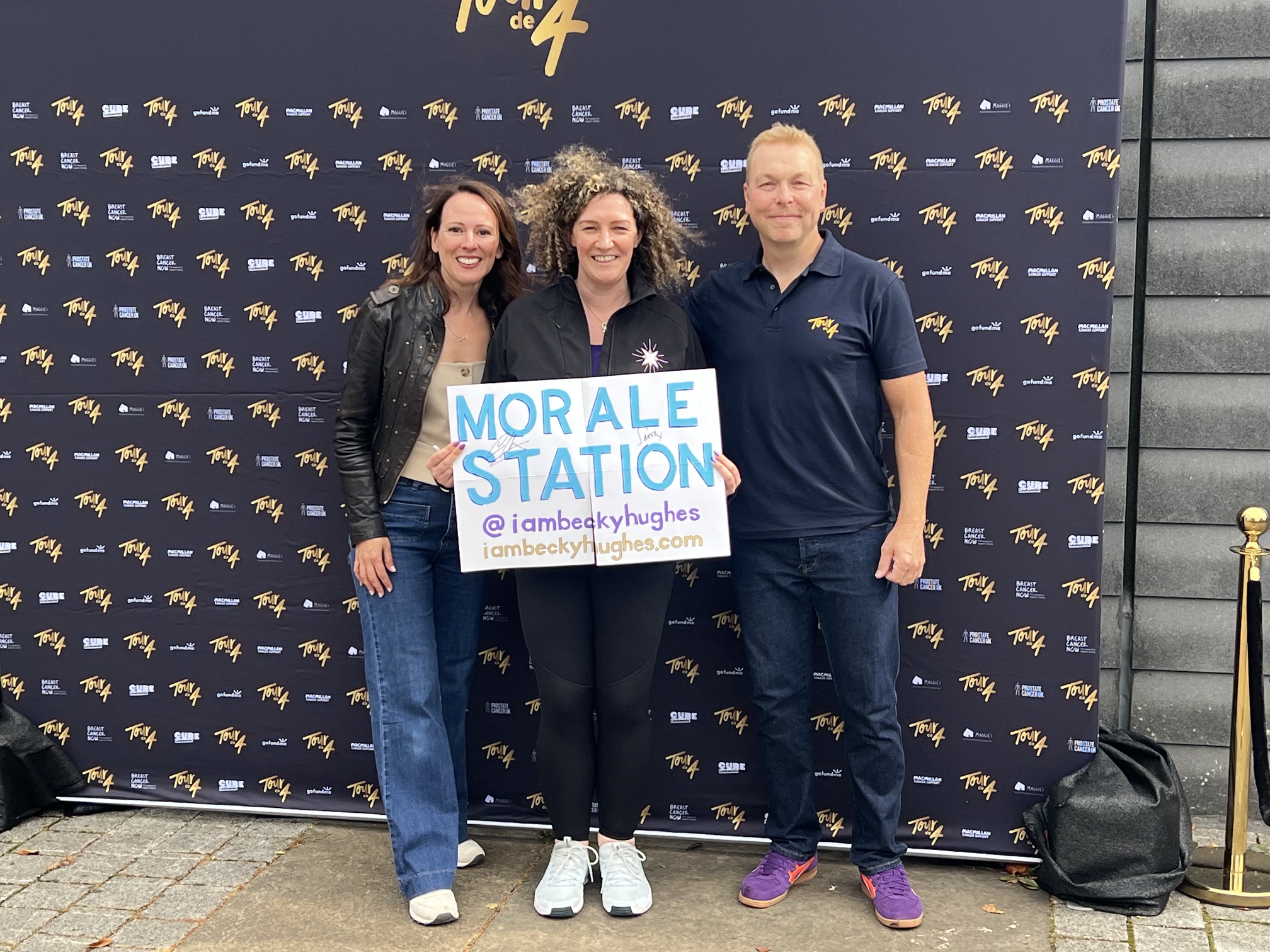 Lady Sarra Hoy, Becky, and Sir Chris Hoy pose with Becky's "Morale Station" sign in front of a Tour de 4 brand wall